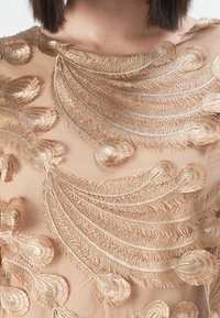 Beige stoff med intrikate gullbroderte bladmønstre, med strukturert detaljering og en glatt, gjennomsiktig finish.
