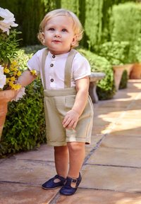 Ljust beige linned shorts med rullade fållar, vit skjorta med mörka knappar, blå läderskor. Barn stående nära blommor och grönska.