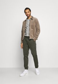 Brown corduroy jacket with a chest pocket, gray striped shirt, green tapered pants, and white sneakers. Simple design, casual fit.