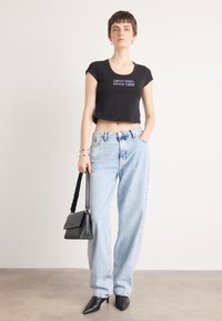 Crna skraćena majica s tiskom "Calvin Klein od 1968.", svijetloplave široke traperice, crna torba i crne cipele s oštrim vrhom.