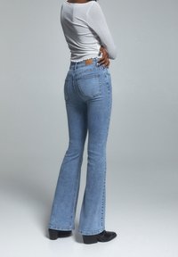Jeans flare denim biru muda dengan saku belakang dan tambalan kulit di pinggang, dipadukan dengan atasan lengan panjang putih yang pas.
