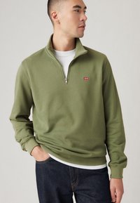 Groene ritsvest van zacht materiaal met een geribde kraag en manchetten, voorzien van een klein rood Levi's-logo op de borst. Gecombineerd met donkere jeans.