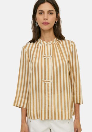 Mujer con una blusa de rayas verticales beige y blanca, con mangas 3/4 y detalles decorativos de botones en la parte delantera.