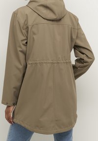 Giacca beige impermeabile con cappuccio, vita sagomata e taglio dritto. Presenta bottoni a scatto ai polsini e una texture liscia.