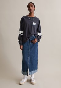 Grijze longsleeve shirt met een "15" graphic en witte gestreepte accenten, gecombineerd met een spijkerrok, witte sneakers en opgerolde sokken.