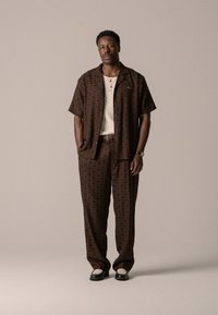 Kurzärmliges braunes Hemd mit Muster und passende Hose im Diamantdesign. Darunter ein helles T-Shirt und schwarze Schuhe.