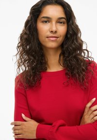 Roter Strickpullover mit Rundhalsausschnitt, langen Ärmeln und gerippten Bündchen. Das Model hat lockige Haare und verschränkt die Arme vor einem neutralen Hintergrund.