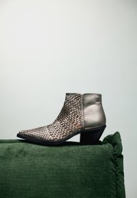 Botín de tobillo de plata trenzada con punta afilada y un tacón grueso negro, que cuenta con un panel lateral metálico liso y un parte superior texturizada.