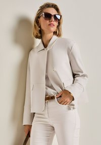 Frau in beiger Jacke und Hose, mit braunem Gürtel, goldenen Ohrringen und Armband, die mit der Hand in der Tasche und Sonnenbrille posiert.