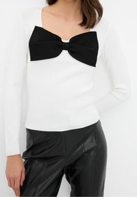 Pull blanc à manches longues avec un grand nœud en tissu noir au niveau du col, associé à un pantalon en similicuir noir. Texture lisse, coupe cintrée.