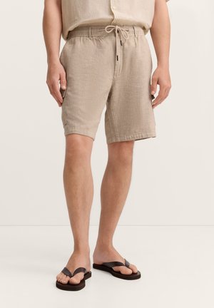 Mann trägt beige Leinen-Shorts mit Kordelzug in der Taille und dunkelbraune Flip-Flops, steht vor einem einfachen Hintergrund.