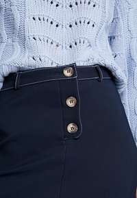 Námořnické kalhoty s vysokým pasem, které mají čtyři velké tmavé knoflíky. Vyznačují se bílým prošíváním podél pasu a hladkým materiálem.