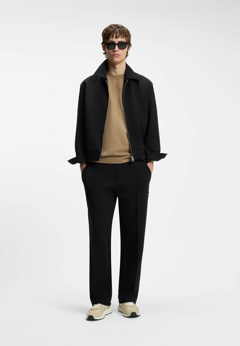 Jeune homme portant une veste noire, un pull beige, un pantalon noir, des baskets beiges et des lunettes de soleil noires, debout les mains dans les poches.