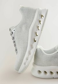 Baskets gris clair avec un dessus en tricot texturé, présentant des semelles blanches élégantes, un col rembourré et des découpes décoratives le long de la base.