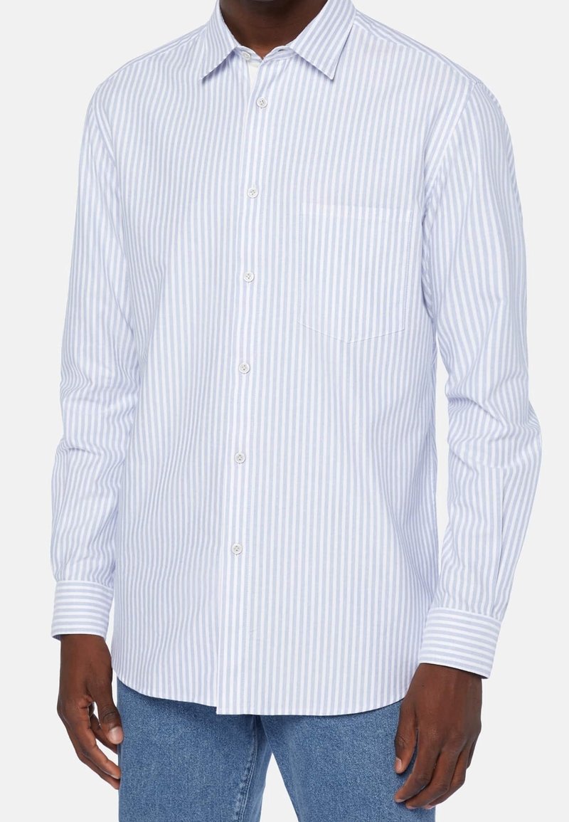 Osoba ve světle modré a bílé pruhované košili s knoflíky stojí na jednobarevném pozadí, s rukama podél těla, v uvolněné póze.