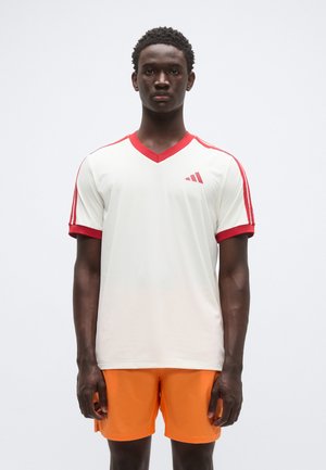 Weißes Sport-T-Shirt mit rotem V-Ausschnitt und roten Akzenten an den Ärmeln, mit roten Seitenstreifen und einem kleinen roten Logo auf der Brust; getragen zu orangen Shorts.