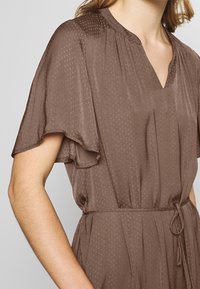 Brown dress featuring a V-neck, short flutter sleeves, and a textured polka dot pattern. Includes a drawstring waist for adjustable fit.