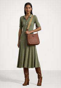 Oliivivihreä polo-tyylinen mekko lyhyillä hihoilla, liehuva helma. Ruskea nahkalaukku kohokuvioidulla logolla ja kuviollisella hihnalla. Polven yli ulottuvat ruskeat saappaat.