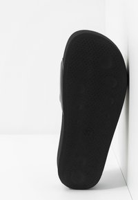 Semelle d'une sandale noire avec un motif texturé antidérapant reposant verticalement contre un mur blanc d'angle.