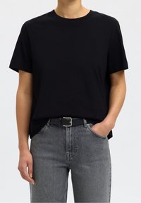 Personne portant un t-shirt noir uni à manches courtes rentré dans un jean gris taille haute avec une ceinture noire, une main dans la poche.