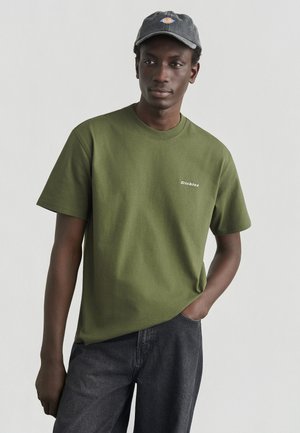 Camiseta de algodón verde oliva con mangas cortas y escote redondo, con el logo blanco de "Dickies" en el pecho, combinada con pantalones gris oscuro.
