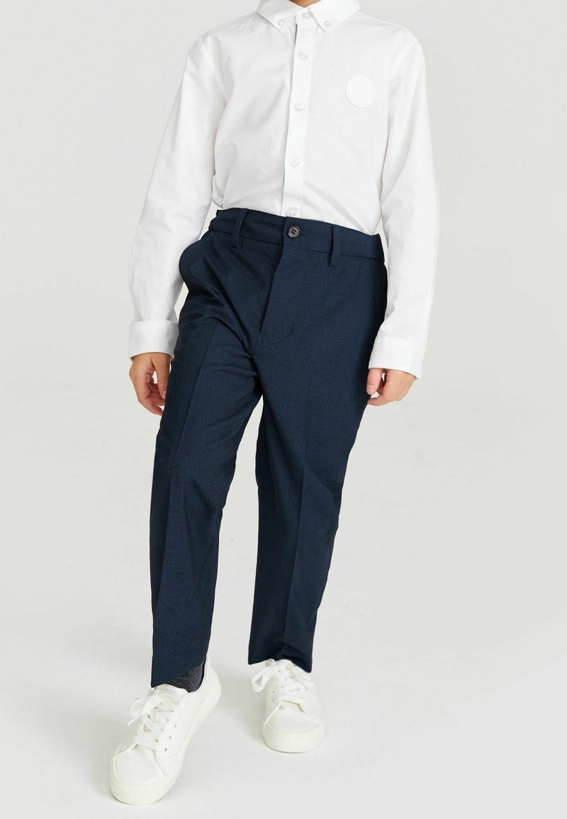 Camicia bianca con bottoni e un piccolo dettaglio circolare, abbinata a pantaloni blu navy con motivo a trama e chiusura con bottoni. Sneakers bianche.