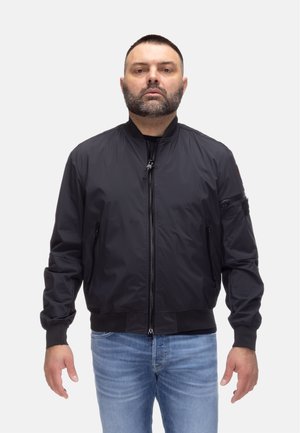 Uomo con barba corta che indossa una giacca bomber nera con zip e jeans azzurri, in piedi davanti a uno sfondo bianco uniforme.