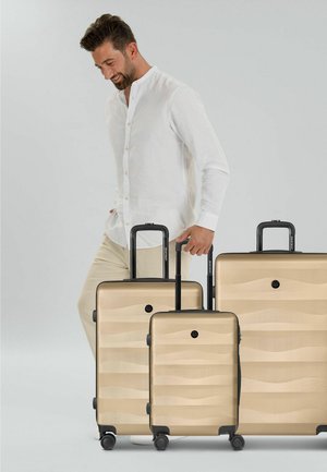 Hombre con camisa blanca y pantalones beige sonriendo mientras camina junto a tres maletas rígidas beige con asas extendidas.