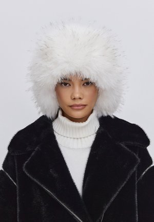 White faux fur hat with a fluffy texture, featuring a rounded shape. Paired with a black shearling coat and a white ribbed turtleneck sweater.