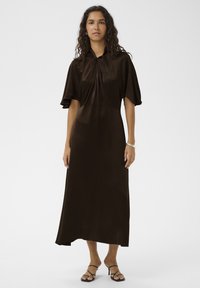 Robe marron mi-longue avec manches courtes, un col, des détails plissés et une texture lisse. Associée à des sandales noires à brides.