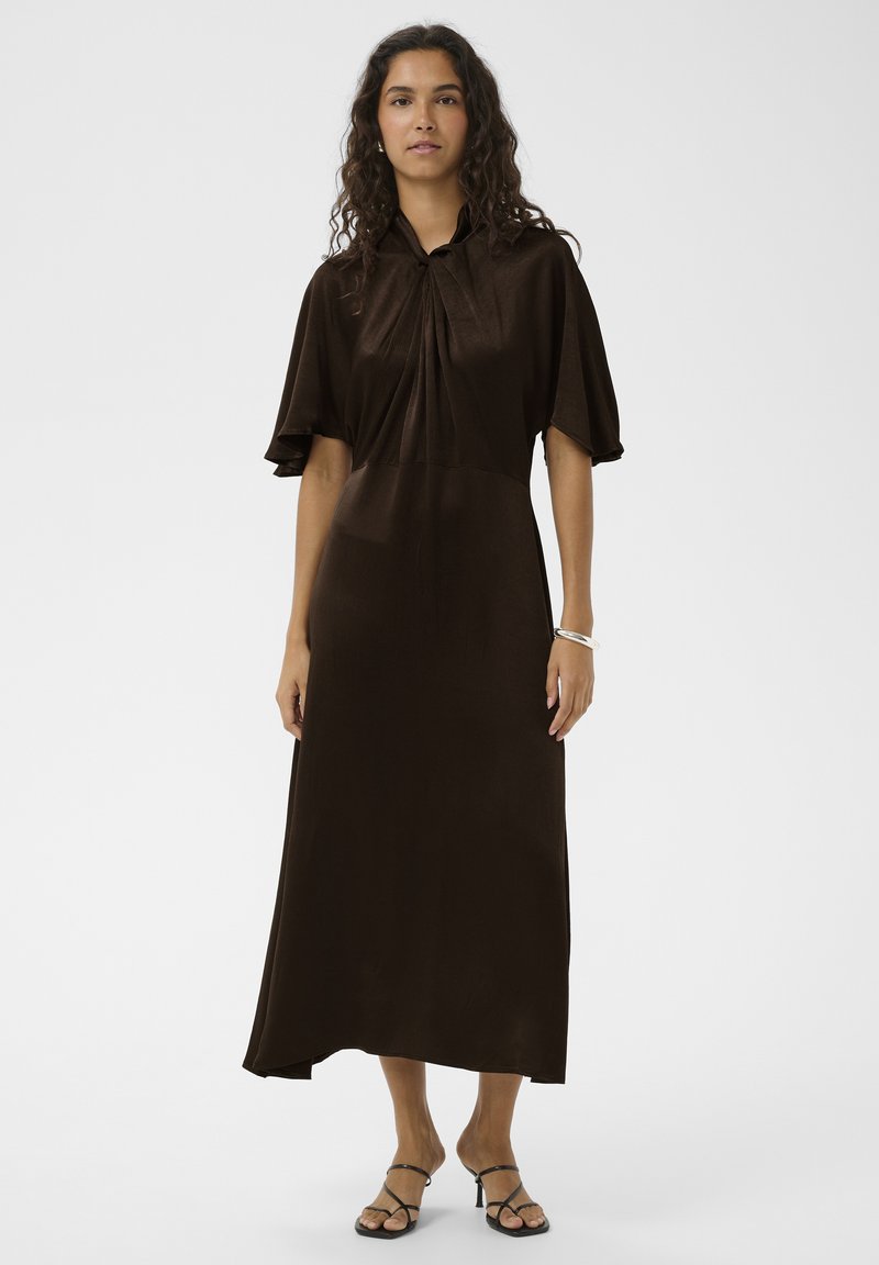Robe marron mi-longue avec manches courtes, un col, des détails plissés et une texture lisse. Associée à des sandales noires à brides.