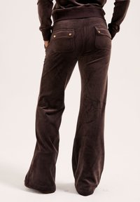 Bruine, uitlopende broek van zacht, gestructureerd materiaal met twee achterzakken met gouden knopen. Soepele afwerking met een aansluitende tailleband.