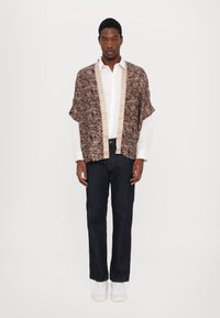 Brunmønstret kimono-stil jakke med beige kanter over en hvid skjorte, kombinert med mørke jeans og hvite sneakers.