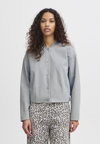 Veste grise coupe courte avec un col rond, des manches longues et une fermeture à boutons. Portée sur un pantalon beige à motif léopard. Texture lisse.