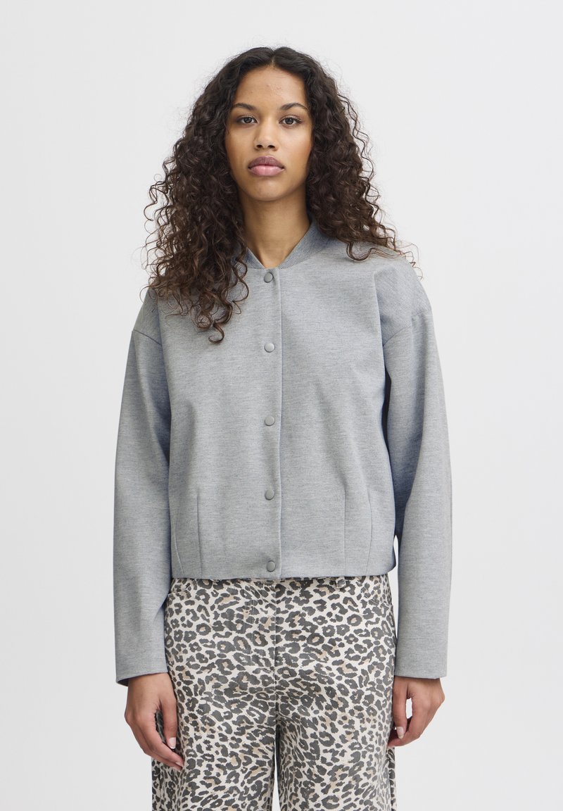 Veste grise coupe courte avec un col rond, des manches longues et une fermeture à boutons. Portée sur un pantalon beige à motif léopard. Texture lisse.