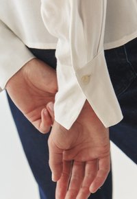 White long-sleeve shirt with a buttoned cuff, smooth texture, and a split hem, worn with dark jeans, hands clasped behind the back.