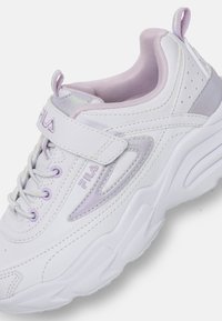 Sneaker Fila blanc et violet clair avec une semelle épaisse, un laçage à l'avant et une sangle à velcro sur un fond uni.