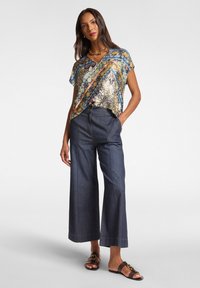 Bunt gemusterte Bluse mit lockerer Passform, kombiniert mit dunkelblauen lässigen Weitbeinen. Braune flache Sandalen mit goldener Schnalle.