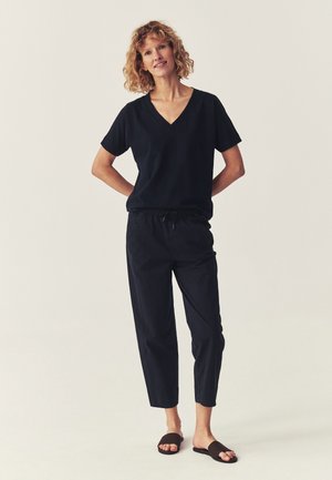 Kobieta o kręconych blond włosach stoi z rękami za plecami, ubrana w czarny top z dekoltem w serek, czarne spodnie z ściągaczem i czarne klapki.