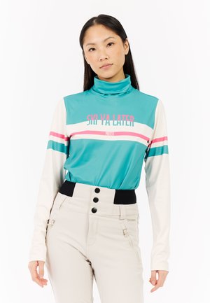 Vrouw in een turquoise en witte lange mouw ski-top met de tekst "SKI YA LATER" en beige skibroek met zwarte knopen.
