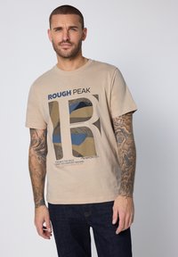 Beige bomulls-t-shirt med tryck som visar blå och bruna former. Texten lyder "ROUGH PEAK" och längst ner finns ett motiverande citat.