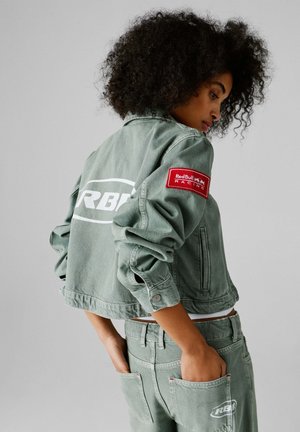 Frau mit lockigem Haar, die eine grüne Denimjacke und passende Hose mit Red Bull Racing-Patches trägt, Hände in den Gesäßtaschen, schaut zur Seite.