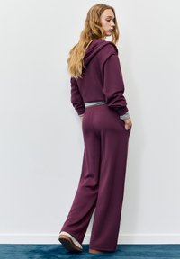 Zestaw do noszenia w domu w ciemnym fioletowym kolorze, zawierający krótki kaptur z szarym obszyciem oraz spodnie o szerokich nogawkach z bocznymi kieszeniami, wykonany z miękkiej tkaniny.