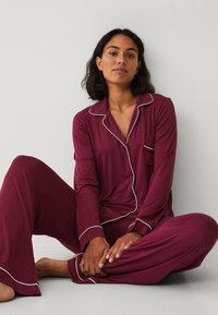 Burgundská pyžamová souprava s bílými lemy; košile na knoflíky s kapsou; volné kalhoty. Měkká látka, dlouhé rukávy, uvolněný střih.