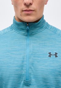 Κοντινό πλάνο ενός ατόμου που φοράει αθλητικό μπλουζάκι με φερμουάρ σε ανοιχτό γαλάζιο χρώμα, με υφή και το λογότυπο Under Armour στο στήθος.