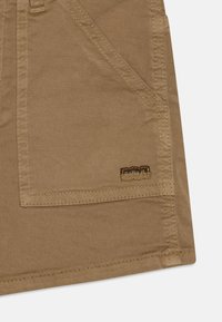Khaki stoff med en glatt tekstur, med en stor lomme og merkelapp. Synlige sømmer langs kantene forbedrer holdbarheten.