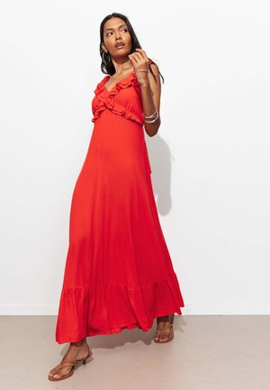 Femme portant une robe maxi rouge sans manches avec des détails à volants, des sandales marron, des boucles d’oreilles anneaux en argent et des bracelets en argent, debout contre un mur blanc.