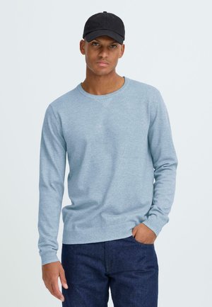 Jonge man met een zwarte pet, lichtblauwe trui en donkeblauwe jeans, staand met één hand in de zak tegen een effen achtergrond.