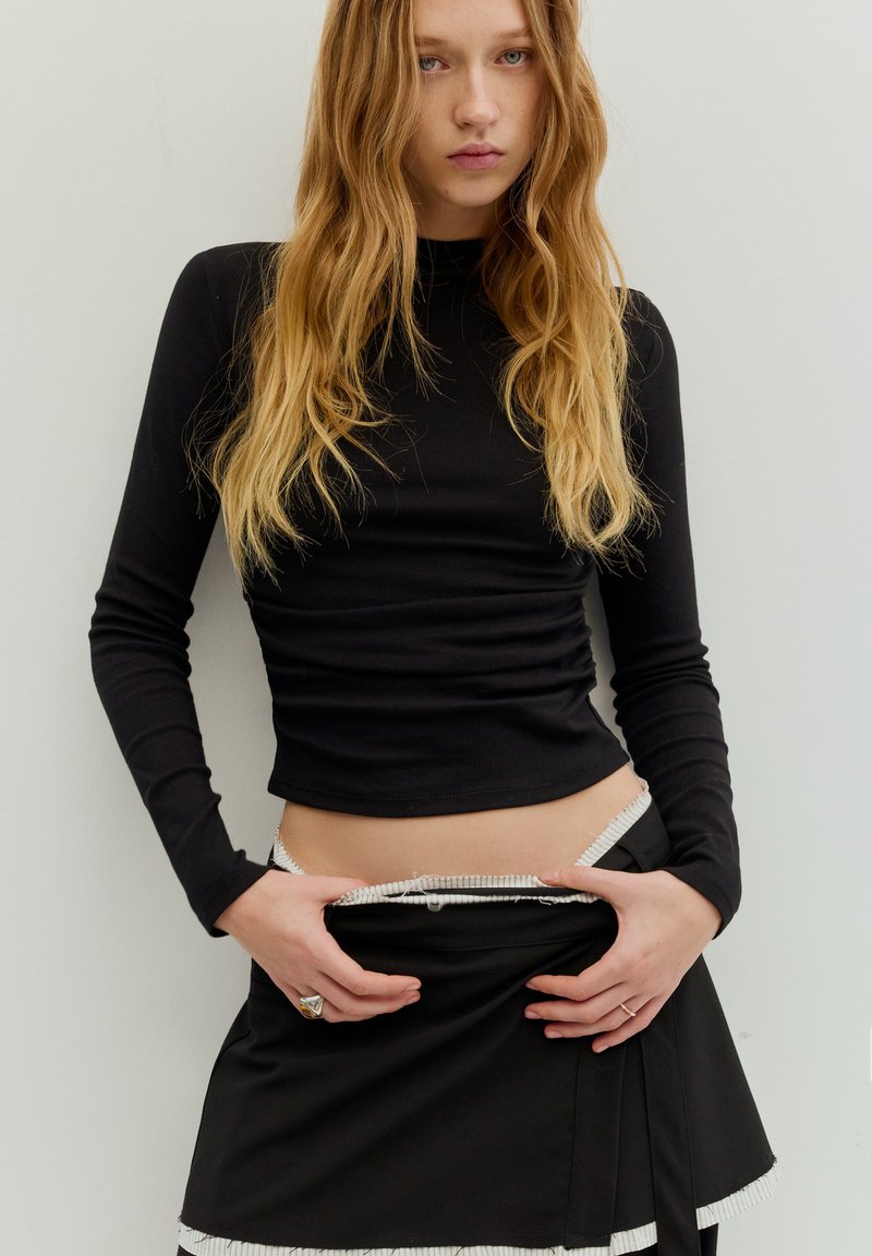 Sort langærmet top med tætsiddende design og tekstureret stof; kombineret med et sort nederdel med hvid kant og plisseret detalje.