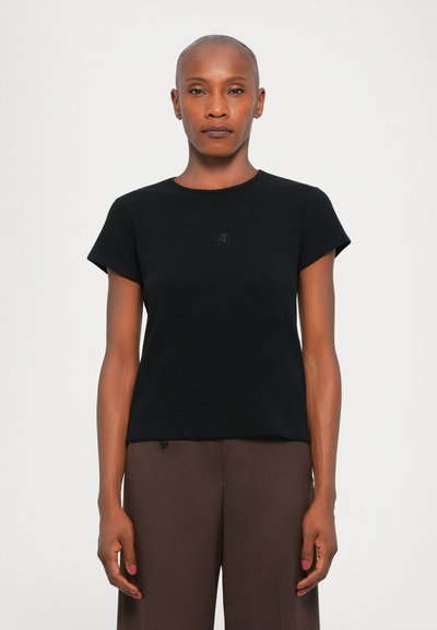 T-shirt noir en coton à manches courtes, col rond et détail de logo subtil sur la poitrine. Porté avec un pantalon marron.